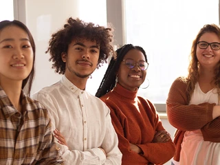 Quatro jovens profissionais de diversas etnias posicionados lado a lado num ambiente de escritório bem iluminado, sorrindo para a câmara de braços cruzados.
